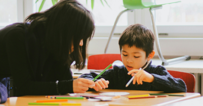 A woman sits at a table helping a child colour