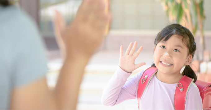 A girl with ponytails and a pink backpack waves goodbye to an adult