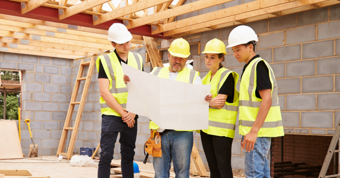 A group of building volunteers in hard hats examine a blueprint at a job site