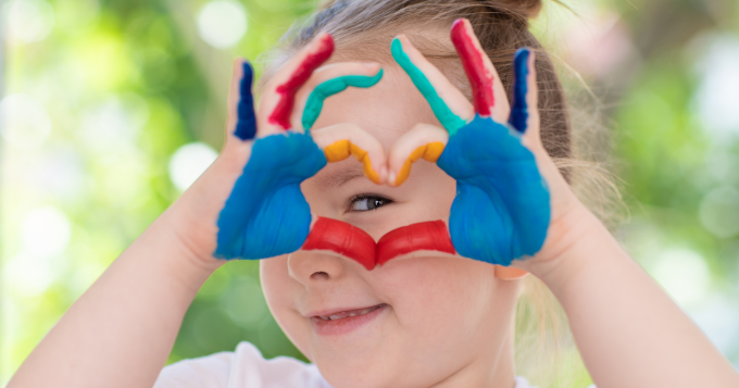 A child with paint on her hands peeks through a heart made with thumbs and fingers