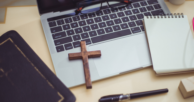 A wooden cross lays on a desk near a keyboard and other office equipment