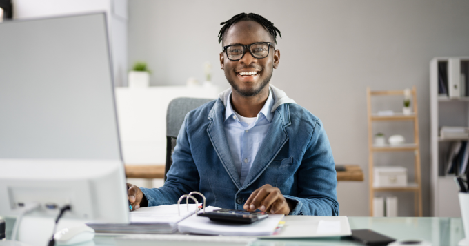 A Black man sits in front of a computer with a calculator smiling