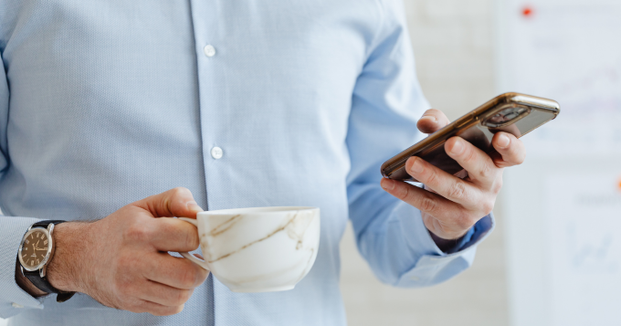 A man in a button down shirt holds a coffee and reads on his phone