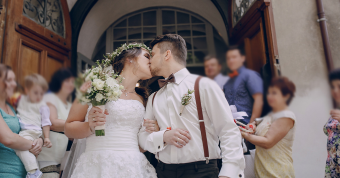 A bride and groom kiss in front of a church door surrounded by family